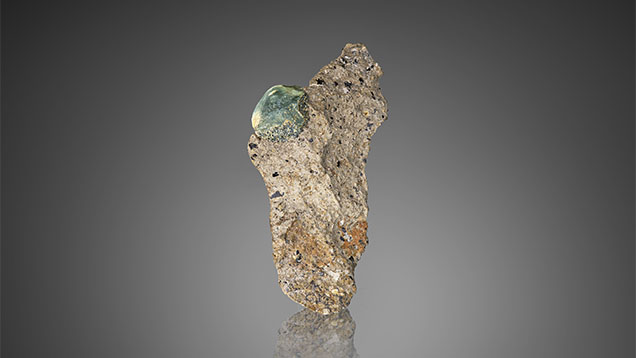 A gem-quality in situ French Bar sill primary sapphire (9 mm long) partially embedded in basaltic trachyandesite rock (sample RK-FBS 001). Photo by Jeff Mason. A gem-quality in situ French Bar sill primary sapphire (9 mm long) partially embedded in basaltic trachyandesite rock (sample RK-FBS 001). Photo by Jeff Mason.