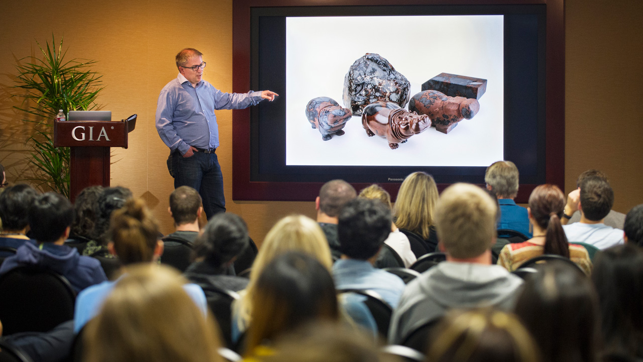 Dreher presented photos of his carvings alongside rough gems to show how he works through each piece to reveal the animal figure. The obsidian hippos pictured on the screen are on display in Carlsbad as part of the “Generations of Mastery” exhibit. Photo by Kevin Schumacher/GIA Dreher presented photos of his carvings alongside rough gems to show how he works through each piece to reveal the animal figure. The obsidian hippos pictured on the screen are on display in Carlsbad as part of the “Generations of Mastery” exhibit. Photo by Kevin Schumacher/GIA