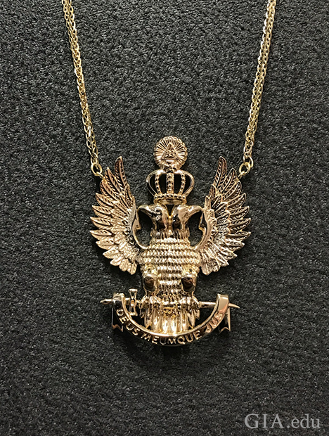 吊坠的造型是两只背靠背坐着、展翅的鹰,它们头上有一顶皇冠。 吊坠的造型是两只背靠背坐着、展翅的鹰,它们头上有一顶皇冠。