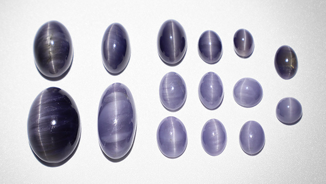 Figure 13. Chatoyant iolite cabochons from Blue Bear. The three largest stones weigh 12.02, 8.52, and 7.24 ct. The remainder weigh between 0.96 to 3.71 ct. Note the variation in color, where lighter-colored cabochons have a higher concentration of acicular inclusions. Photo by Philippe Belley. Figure 13. Chatoyant iolite cabochons from Blue Bear. The three largest stones weigh 12.02, 8.52, and 7.24 ct. The remainder weigh between 0.96 to 3.71 ct. Note the variation in color, where lighter-colored cabochons have a higher concentration of acicular inclusions. Photo by Philippe Belley.