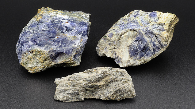 Figure 9. Representative cordierite (top left), quartz with cordierite (top right), and gedrite (bottom) from Blue Bear. As is common in metamorphic gem deposits, only a small fraction of the mineral occurs in gem quality. The gedrite fragment measures 65 mm across. Photo by Philippe Belley. Figure 9. Representative cordierite (top left), quartz with cordierite (top right), and gedrite (bottom) from Blue Bear. As is common in metamorphic gem deposits, only a small fraction of the mineral occurs in gem quality. The gedrite fragment measures 65 mm across. Photo by Philippe Belley.