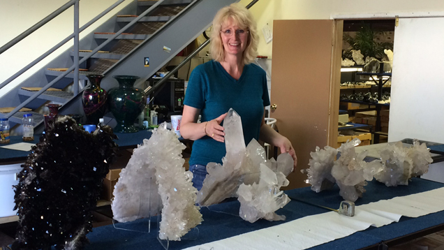 Terri Ottaway stands behind a table with several very large quartz crystal specimens are lined up on it. Some of them come up to her waist height. Terri Ottaway stands behind a table with several very large quartz crystal specimens are lined up on it. Some of them come up to her waist height.