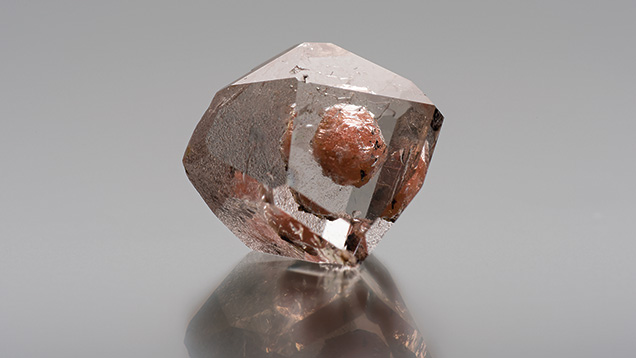 Pinkish orange inclusion in topaz.. Figure 1. A pinkish orange inclusion dominates the interior of this 20.58 ct unpolished topaz crystal from Pakistan. Photograph by Kevin Schumacher.. Small trigons and a rough surface texture seen on a chondrodite crystal.