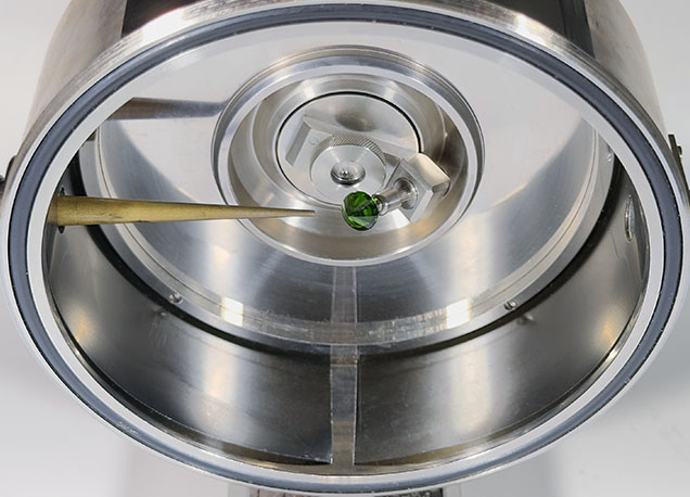 A close-up view of a metallic chamber with a green object at the center, featuring a tool pointing towards it. A close-up view of a metallic chamber with a green object at the center, featuring a tool pointing towards it.