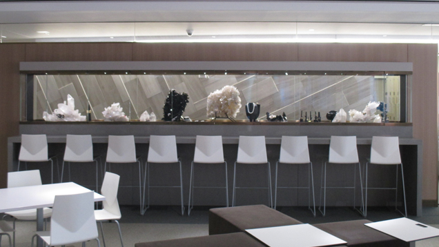 A view of the dramatic long, narrow exhibit case filled with GIA Museum pieces. The case is a feature wall in the student commons area filled with tables and chairs. A view of the dramatic long, narrow exhibit case filled with GIA Museum pieces. The case is a feature wall in the student commons area filled with tables and chairs.